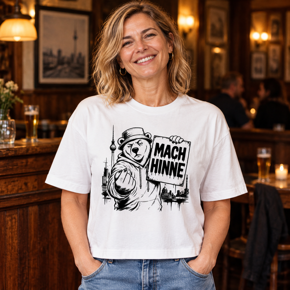 Lachende Frau in einer gemütlichen Berliner Kneipe trägt ein weißes, locker geschnittenes Damen-T-Shirt mit schwarzem Berliner-Bär-Motiv, dem Schriftzug „MACH HINNE“, Berliner Skyline und Fernsehturm.
