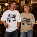 Ein lachender Mann und eine lachende „Lachender Mann und lachende Frau, beide etwa 45 bis 60 Jahre alt, in einer authentischen Berliner Kneipe. Sie tragen locker geschnittene Oversize-T-Shirts in Weiß und Steingrau mit schwarzem Berliner-Bär-Motiv, dem Schriftzug „Mach hinne“ und Berliner Skyline mit Fernsehturm.
