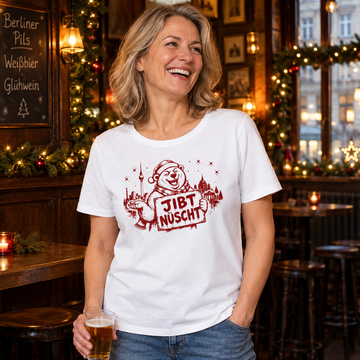 Lachende Frau zwischen 45 und 60 Jahren in einer weihnachtlich geschmückten Berliner Kneipe mit weißem Bio-Damen-T-Shirt und rotem „JIBT NÜSCHT“-Motiv mit Weihnachtsbär und Berliner Skyline.