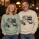 Lachender Mann und lachende Frau in einer gemütlichen Berliner Kneipe tragen Unisex-Sweater in hellem grau-grün und Beige mit schwarzem „Mach hinne“-Motiv: Berliner Bär, Berliner Skyline und Fernsehturm.“