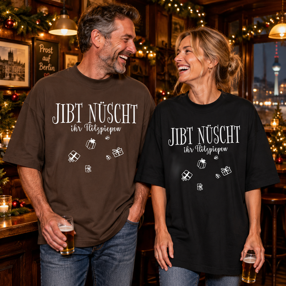 Lachendes Paar in einer weihnachtlich geschmückten Berliner Kneipe: Mann im braunen und Frau im schwarzen Oversize-Bio-T-Shirt mit weißem „JIBT NÜSCHT, ihr Flitzpiepen“-Weihnachtsmotiv und Geschenk-Illustrationen.