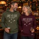 Lachendes Paar in einer weihnachtlich geschmückten Berliner Kneipe: Mann im khaki-grünen und Frau im bordeauxfarbenen  Bio-Hoodie mit weißem „JIBT NÜSCHT, ihr Flitzpiepen“-Weihnachtsmotiv und Geschenk-Illustrationen.
