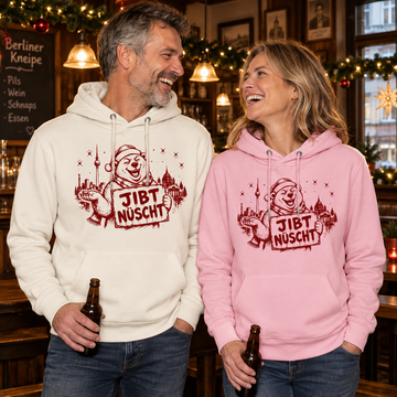 Lachendes europäisches Paar zwischen 45 und 60 Jahren in einer weihnachtlich geschmückten Berliner Kneipe: Die Frau trägt einen rosa, der Mann einen naturweißen Unisex Bio-Hoodie mit rotem „JIBT NÜSCHT“-Weihnachtsmotiv, Berliner Skyline und Weihnachtsbär.