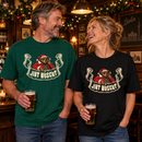 Lachendes Paar zwischen 45 und 60 Jahren in einer weihnachtlich geschmückten Berliner Kneipe: Der Mann trägt ein dunkelgrünes, die Frau ein schwarzes Relaxed-Fit-Bio-T-Shirt mit Santa-Bär-Motiv und dem Schriftzug „JIBT NÜSCHT“.