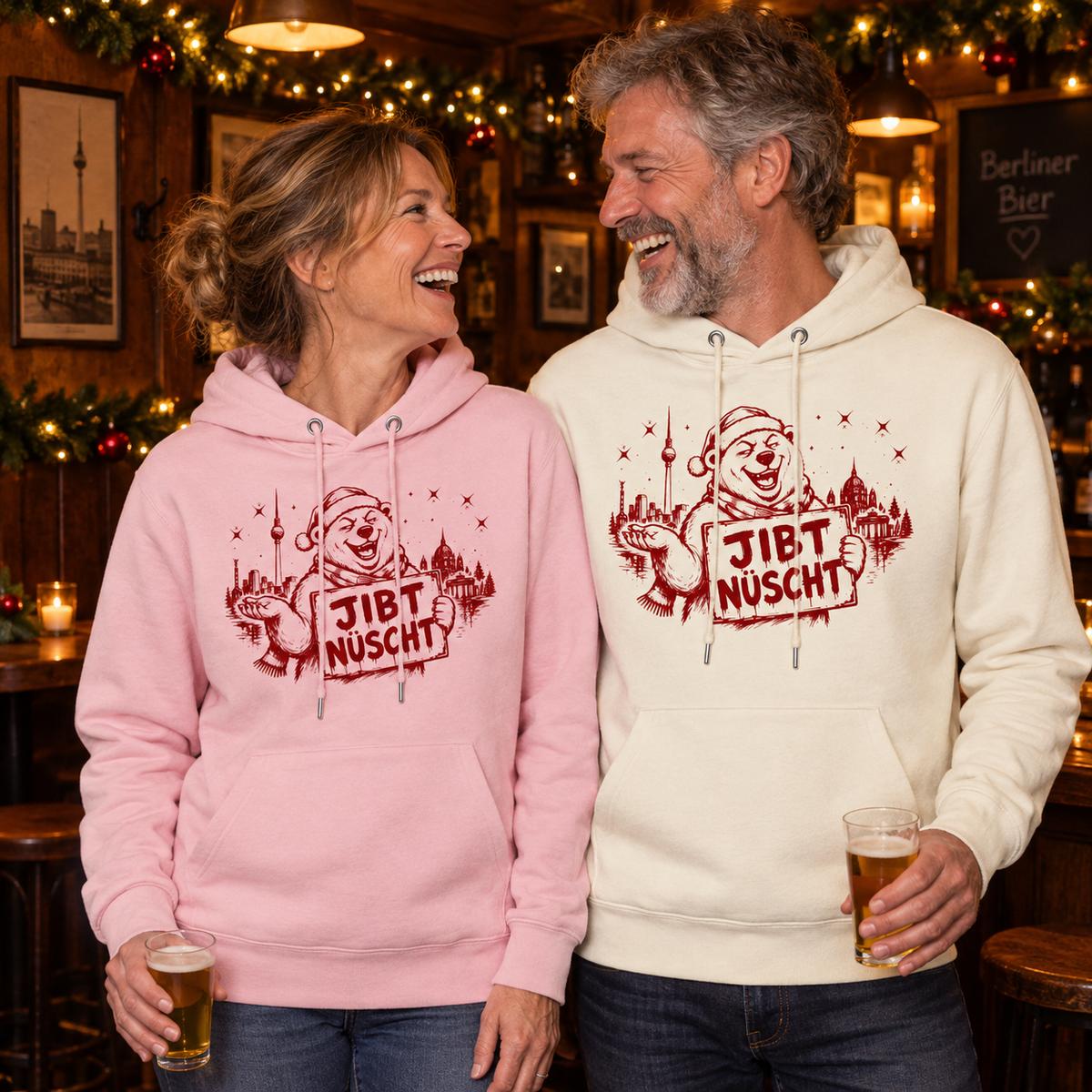 Lachendes europäisches Paar zwischen 45 und 60 Jahren in einer weihnachtlich geschmückten Berliner Kneipe: Die Frau trägt einen rosa, der Mann einen naturweißen Unisex Bio-Hoodie mit rotem „JIBT NÜSCHT“-Weihnachtsmotiv, Berliner Skyline und Weihnachtsbär.