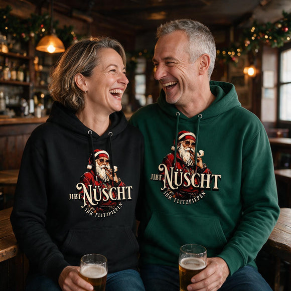 Lachendes Paar in einer weihnachtlich geschmückten Berliner Kneipe: Mann im grünen und Frau im schwarzen Unisex-Bio-Hoodie mit Berliner Gangster-Weihnachtsmotiv, rauchendem Weihnachtsmann und dem Schriftzug „JIBT NÜSCHT, ihr Flitzpiepen“.
